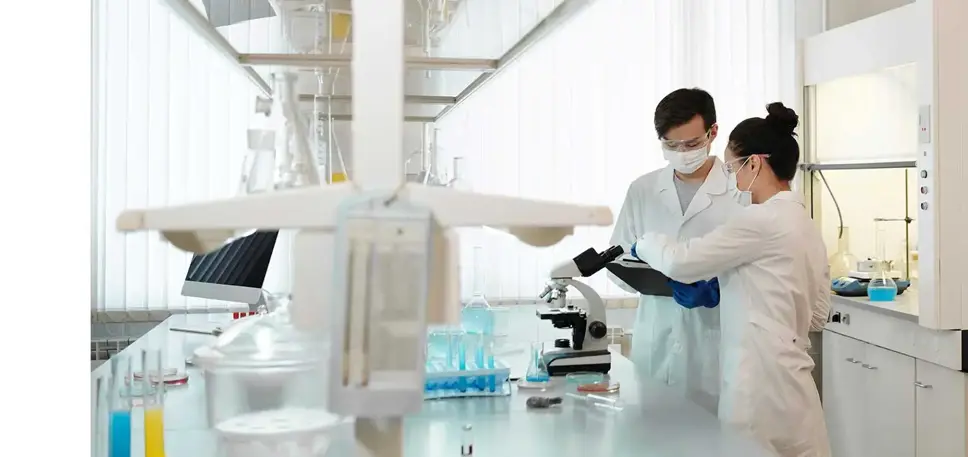 Two scientists discussing are standing in a lab in front of a microscope on the bench.