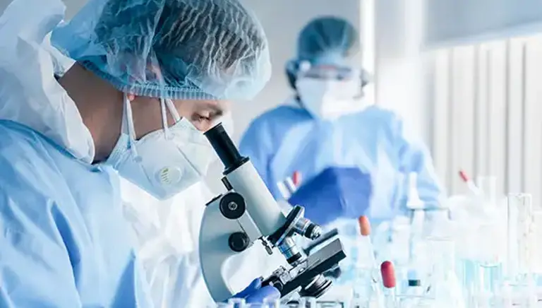 Two masked scientists in a lab, the one in the background takes note with the closer one looks into a microscope