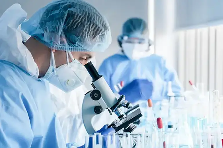 Two masked scientists in a lab, the one in the background takes note with the closer one looks into a microscope