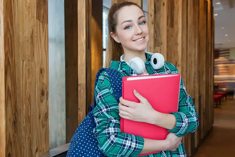 Young smiling college student, with backpack and headphones, holds a ring binder in her arms