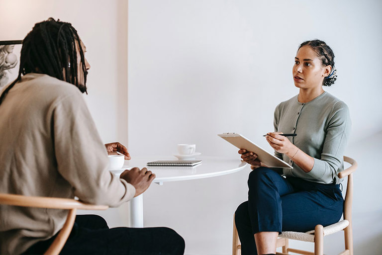 Two mixed raced people are sat drinking tea, in a office, while one seems to be interviewing the other. 