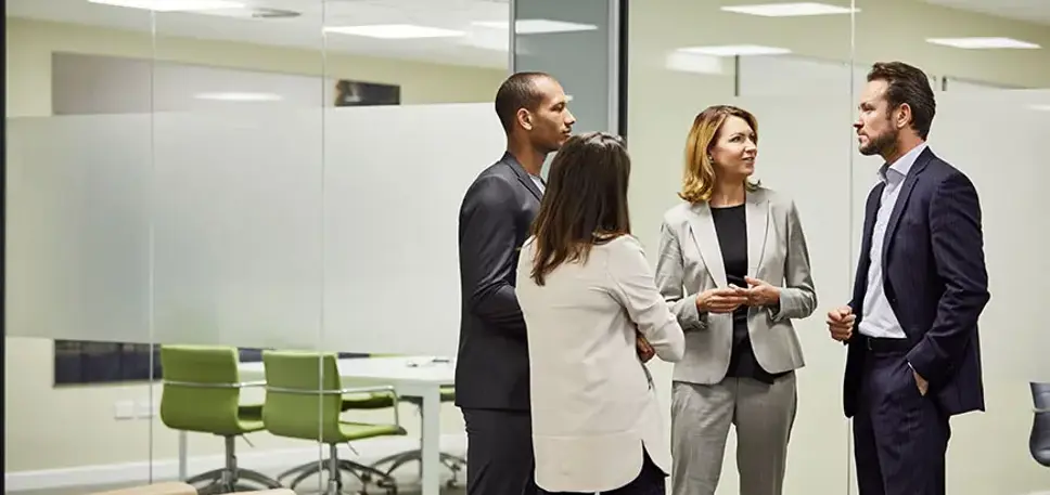 Four office workers stand talking in front of a set of glass meeting rooms