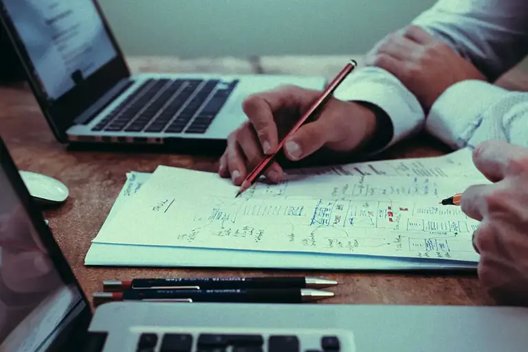 Close-up of the hands of two office workers writing notes on a desk between to open laptops