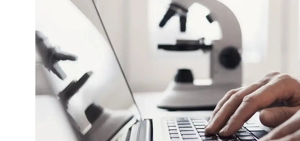 Close-up of hands typing on a laptop, with a microscope in the background, being reflected on the screen