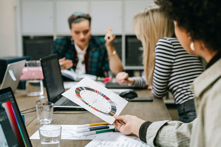 Three women discuss coloured charts from a survey around a business meeting table. 